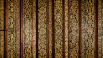 Intricate vertical patterns cover the surface, featuring ornate geometric and floral motifs in gold and brown hues. The repeating designs suggest a traditional or historical style, possibly inspired by Middle Eastern or Asian art. The arrangement appears symmetrical and is executed with fine detail and precision.
