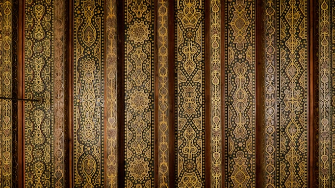 Intricate vertical patterns cover the surface, featuring ornate geometric and floral motifs in gold and brown hues. The repeating designs suggest a traditional or historical style, possibly inspired by Middle Eastern or Asian art. The arrangement appears symmetrical and is executed with fine detail and precision.