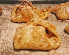 Golden-brown pastry squares with a flaky texture, lightly dusted with powdered sugar. The pastries have a rustic appearance with some folds and creases.