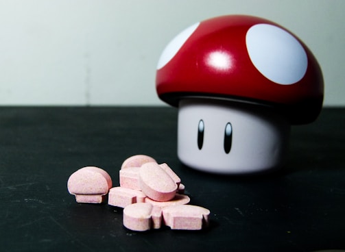 A toy resembling the iconic mushroom from a popular video game is placed next to a small pile of pink, mushroom-shaped tablets on a flat black surface. The toy has a red cap with white spots and eyes on the stem, mimicking a cartoonish, playful design.