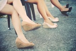 Three pairs of legs are visible seated on chairs, wearing wedge heels and sandals. The ground is a grey concrete surface, and the style of the shoes suggests a casual or semi-formal setting.