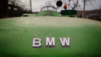 A close-up of a green vehicle's hood with visible wear and tear. The letters 'BMW' are prominently displayed in silver, centered in the image. In the background, blurred objects suggest an outdoor setting, possibly a rural or farm area.