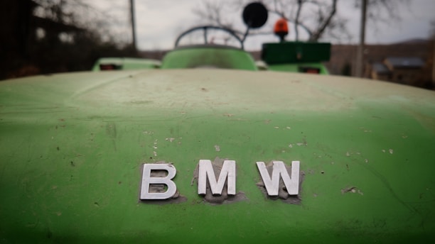 A close-up of a green vehicle's hood with visible wear and tear. The letters 'BMW' are prominently displayed in silver, centered in the image. In the background, blurred objects suggest an outdoor setting, possibly a rural or farm area.