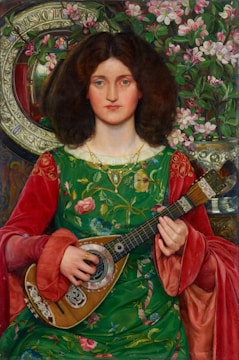 A woman with long, curly brown hair is dressed in an ornate, green floral gown with red sleeves. She holds a string instrument resembling a mandolin or lute. In the background, there is an intricate floral pattern and decorative items including a round mirror with decorative elements and a large vase of pink flowers.