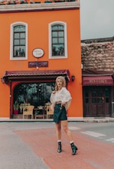A person is standing in front of a vibrant orange building with two large windows and a sign labeled 'nevi cafe'. The individual is dressed in a white blouse, dark skirt, and black boots, posing with one hand on the hip. The setting includes outdoor seating with wicker chairs and plants visible through the window.