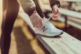 A person is tying the laces of a white athletic shoe with a noticeable logo, sitting on a wooden bench. The lighting suggests a warm and possibly outdoor setting.