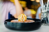 A dish sits on a table featuring a dessert with a scoop of vanilla ice cream drizzled with caramel sauce on top of waffles. The background includes blurred figures and warm lighting, contributing to a cozy atmosphere.