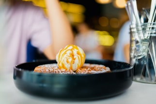 A dish sits on a table featuring a dessert with a scoop of vanilla ice cream drizzled with caramel sauce on top of waffles. The background includes blurred figures and warm lighting, contributing to a cozy atmosphere.