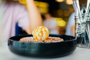 A dish sits on a table featuring a dessert with a scoop of vanilla ice cream drizzled with caramel sauce on top of waffles. The background includes blurred figures and warm lighting, contributing to a cozy atmosphere.