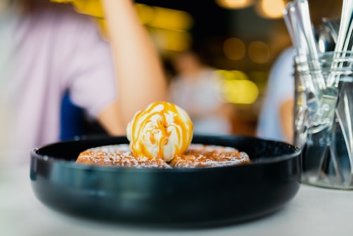 A dish sits on a table featuring a dessert with a scoop of vanilla ice cream drizzled with caramel sauce on top of waffles. The background includes blurred figures and warm lighting, contributing to a cozy atmosphere.