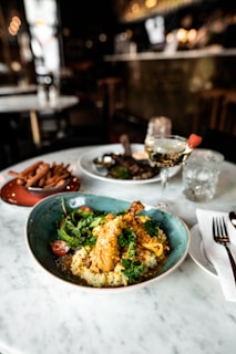 A plate of gourmet food with a chicken drumstick on a bed of creamy risotto, garnished with fresh herbs and vegetables, sits on a marble table in a dimly lit restaurant setting. A glass of white wine and cutlery are also present.