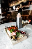 A charcuterie board on a marble table holding a variety of meats, cheeses, olives, and grapes, accompanied by a glass of champagne with a strawberry garnish and a bottle in an ice bucket. The background is a blurred view of a bar with dim lighting.