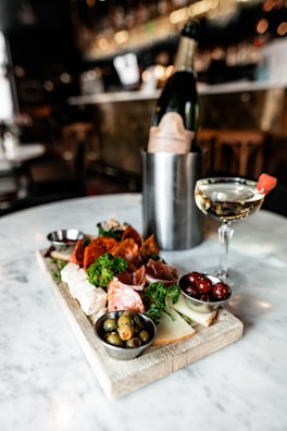 A charcuterie board on a marble table holding a variety of meats, cheeses, olives, and grapes, accompanied by a glass of champagne with a strawberry garnish and a bottle in an ice bucket. The background is a blurred view of a bar with dim lighting.