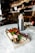 A charcuterie board on a marble table holding a variety of meats, cheeses, olives, and grapes, accompanied by a glass of champagne with a strawberry garnish and a bottle in an ice bucket. The background is a blurred view of a bar with dim lighting.