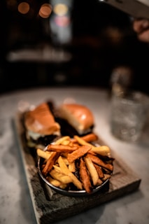 A plate of fries is served next to two burgers on a wooden board. The scene appears to be in a dimly lit setting, likely a restaurant or a cafe, with a bokeh effect visible in the background.