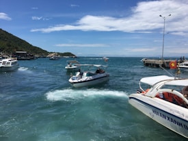 Several boats are navigating in a clear, blue sea near a small dock. The sky is partly cloudy, with a hill covered in greenery in the background. A few people can be seen on the dock and on the boats themselves.