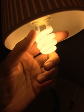 A hand is holding a compact fluorescent light bulb under a lamp shade, illuminating the surrounding area with a warm glow.