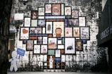 A wall mural composed of numerous colorful tiles, each featuring a different artwork, cartoon character, or illustration. The arrangement forms a tree-like shape against an aged stone wall. On the left, a cutout figure holding a blue sign is visible.