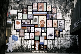 A wall mural composed of numerous colorful tiles, each featuring a different artwork, cartoon character, or illustration. The arrangement forms a tree-like shape against an aged stone wall. On the left, a cutout figure holding a blue sign is visible.