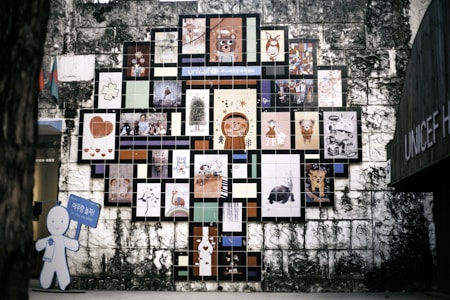 A wall mural composed of numerous colorful tiles, each featuring a different artwork, cartoon character, or illustration. The arrangement forms a tree-like shape against an aged stone wall. On the left, a cutout figure holding a blue sign is visible.