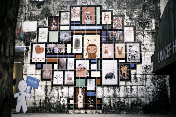 A wall mural composed of numerous colorful tiles, each featuring a different artwork, cartoon character, or illustration. The arrangement forms a tree-like shape against an aged stone wall. On the left, a cutout figure holding a blue sign is visible.