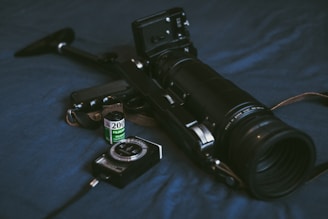 A vintage camera with a large telephoto lens lies on a dark surface accompanied by a roll of Fujicolor film and a light meter. The camera body appears worn, indicating frequent use, and the film roll is positioned upright next to it.