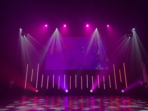 A dimly lit stage features vibrant purple and pink lighting emanating from spotlights and LED bars. The backdrop displays abstract patterns, while the floor reflects circular and linear light patterns, creating an energetic and dynamic visual atmosphere.