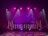 A dimly lit stage features vibrant purple and pink lighting emanating from spotlights and LED bars. The backdrop displays abstract patterns, while the floor reflects circular and linear light patterns, creating an energetic and dynamic visual atmosphere.