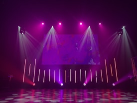 A dimly lit stage features vibrant purple and pink lighting emanating from spotlights and LED bars. The backdrop displays abstract patterns, while the floor reflects circular and linear light patterns, creating an energetic and dynamic visual atmosphere.
