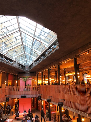 A spacious indoor area with a large glass ceiling allowing natural light to illuminate the space. The architecture features industrial elements like steel beams and exposed brick. The space is populated with people socializing and engaging in leisure activities. Warm lighting and wooden railings add a cozy and inviting atmosphere.