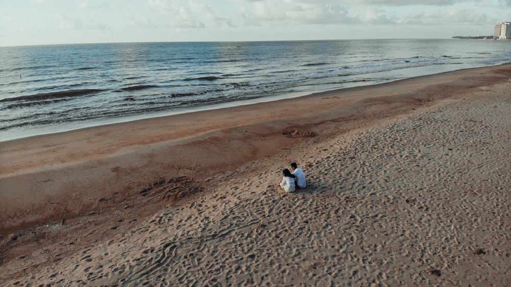 two people sits on sand beside seashore during daytime