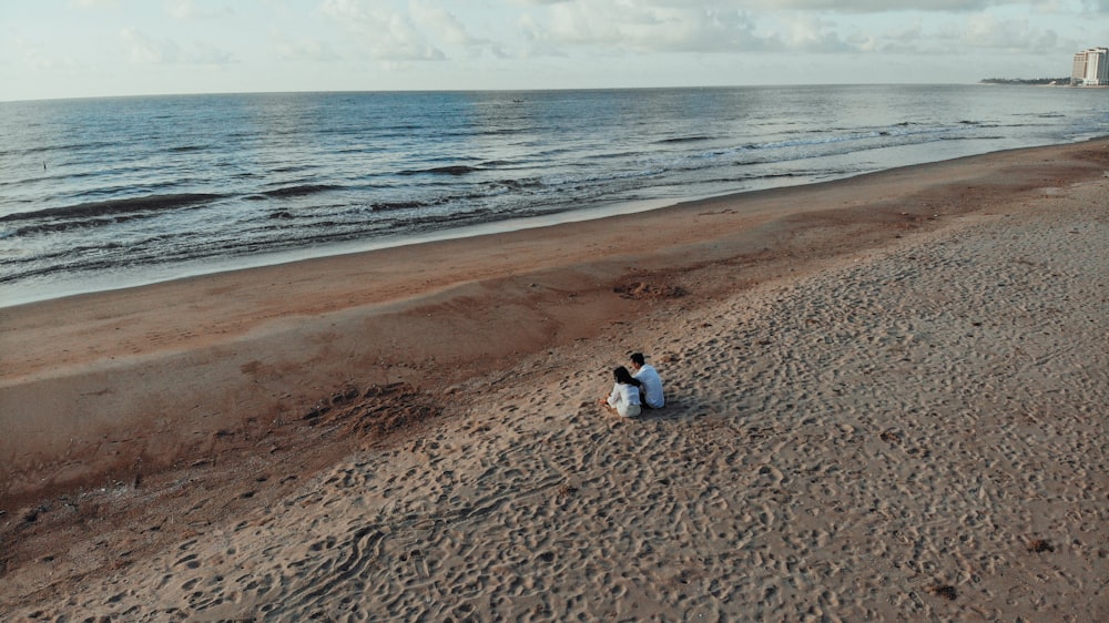 duas pessoas sentam-se na areia ao lado da costa durante o dia