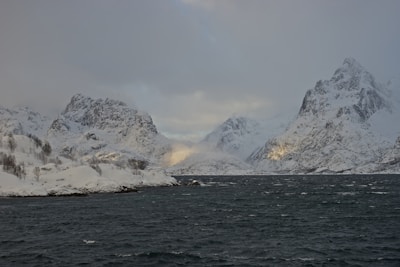 Snow-covered mountains rise majestically on either side of a dark, choppy sea. The rugged peaks are partially shrouded in mist and clouds, adding an air of mystery. Subtle patches of sunlight break through, casting a gentle glow on the mountains.