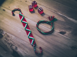 A wooden surface displays three red dice with white dots, a woven friendship bracelet featuring geometric patterns in red, green, and white, and a braided bracelet in matching colors with tassels.