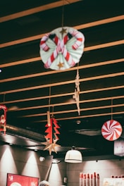 Festive decorations hang from a wooden ceiling, including a candy cane wreath, a red and white peppermint swirl, a red and green tree, and gold star ornaments. The setting appears to be an indoor space with warm lighting, cups stacked on the counter, and a person wearing head protection partially visible.