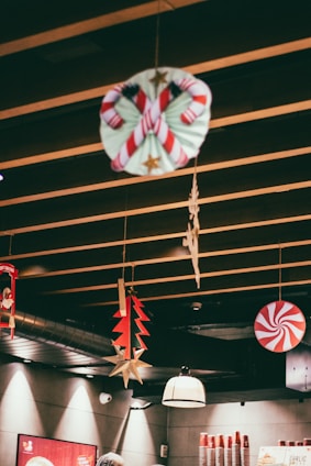 Festive decorations hang from a wooden ceiling, including a candy cane wreath, a red and white peppermint swirl, a red and green tree, and gold star ornaments. The setting appears to be an indoor space with warm lighting, cups stacked on the counter, and a person wearing head protection partially visible.