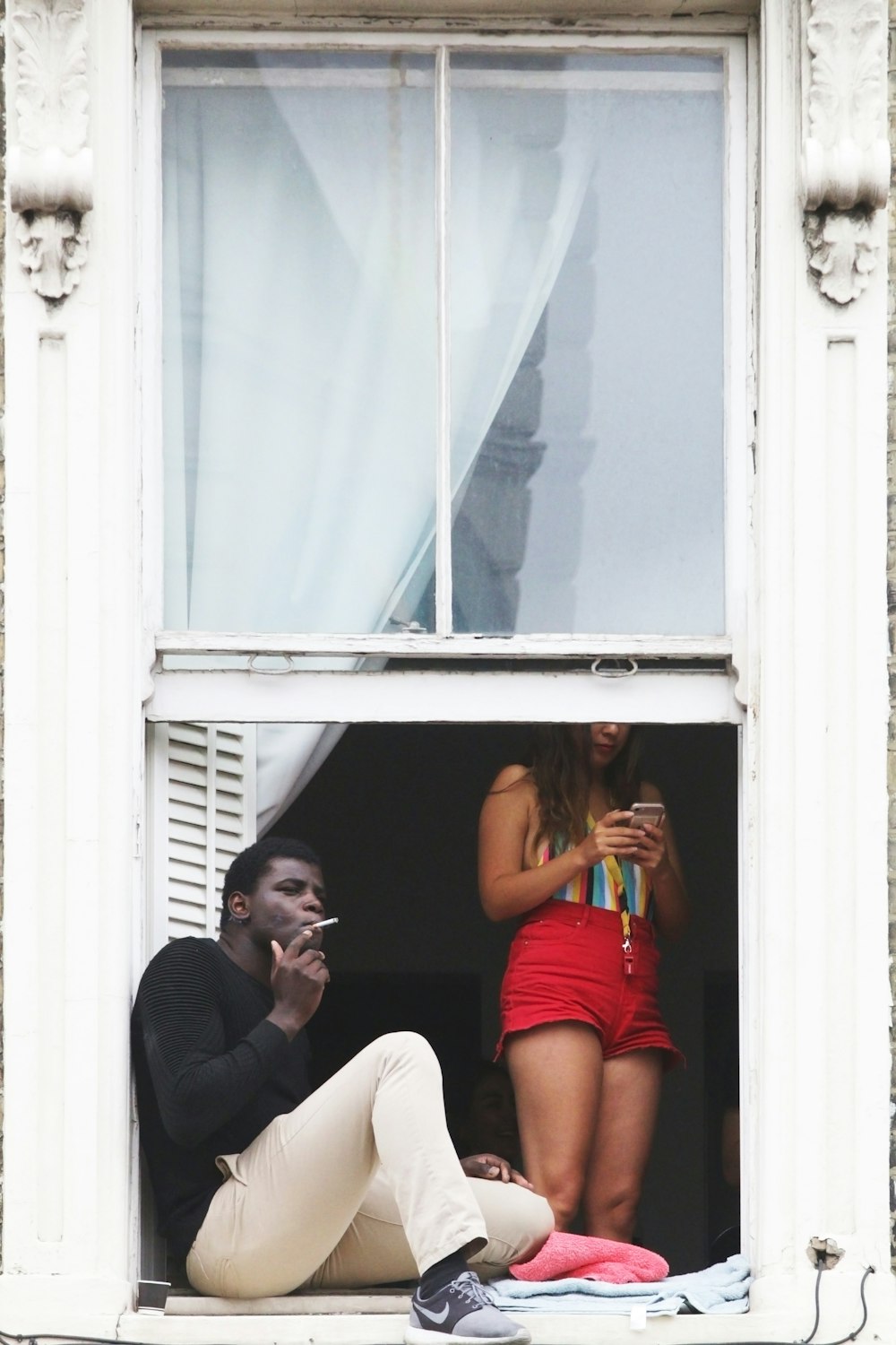 man wearing black crew-neck sweatshirt smoking cigarette stick while sitting near open window near woman using phone standing