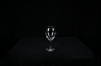 A clear wine glass sits centered on a dark, textured surface, surrounded by a black background. The glass is empty, and its contours are sharply highlighted against the stark backdrop.