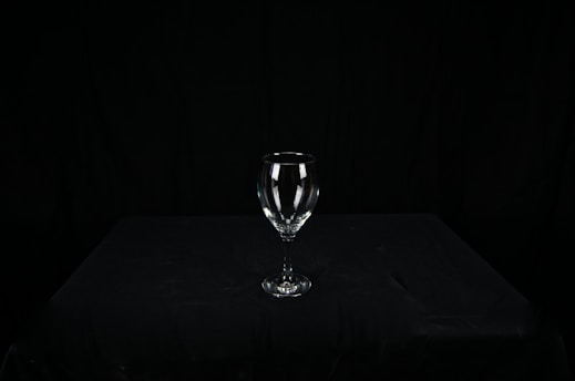 A clear wine glass sits centered on a dark, textured surface, surrounded by a black background. The glass is empty, and its contours are sharply highlighted against the stark backdrop.