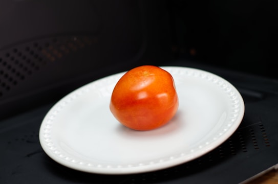 A single ripe tomato is placed on a white ceramic plate with a beaded rim. The background is dark, possibly the inside of a microwave or oven.