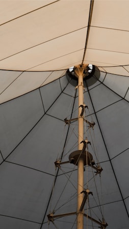 A geometric structure with a central pole surrounded by tension cables, supporting a large fabric canopy that forms a pointed shape with triangular sections.