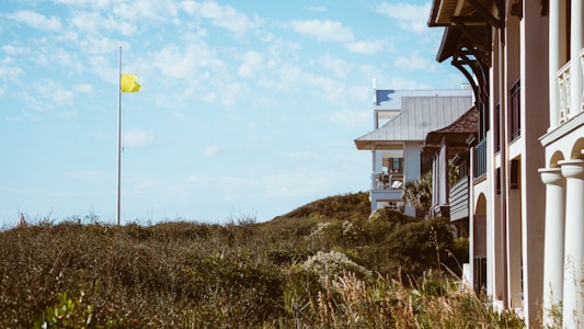 A coastal scene featuring residential buildings with wooden and metal elements on the right side, surrounded by lush greenery. A yellow flag is seen on a tall pole in the middle of the landscape, with a clear blue sky and scattered white clouds overhead.