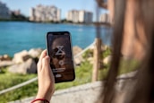 A person holds a smartphone displaying a relaxation application while standing near a body of water with buildings in the background. The scene is lit by natural daylight.