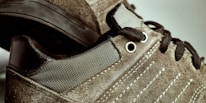 Close-up image of suede sneakers featuring detailed stitching and metal eyelets. The shoes have black laces and textured fabric, with a contrast between the suede and smooth materials.