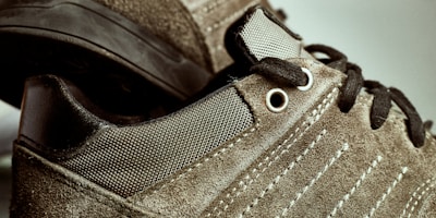 Close-up image of suede sneakers featuring detailed stitching and metal eyelets. The shoes have black laces and textured fabric, with a contrast between the suede and smooth materials.