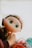 A large-eyed doll with an expression of curiosity is being held by the hand of a child. Its face has freckles, and it has long, wavy hair. Surrounding the doll are blurred items, including a partially visible pair of colorful glasses.