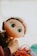 A large-eyed doll with an expression of curiosity is being held by the hand of a child. Its face has freckles, and it has long, wavy hair. Surrounding the doll are blurred items, including a partially visible pair of colorful glasses.