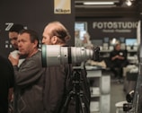 A large camera lens on a tripod is displayed prominently in the foreground with several people interacting in the background. Logos from Nikon and Fujinon are visible, suggesting a photography-related event or exhibition. The setting appears to be indoors, possibly a photo studio or a camera store.