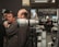 A large camera lens on a tripod is displayed prominently in the foreground with several people interacting in the background. Logos from Nikon and Fujinon are visible, suggesting a photography-related event or exhibition. The setting appears to be indoors, possibly a photo studio or a camera store.