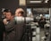 A large camera lens on a tripod is displayed prominently in the foreground with several people interacting in the background. Logos from Nikon and Fujinon are visible, suggesting a photography-related event or exhibition. The setting appears to be indoors, possibly a photo studio or a camera store.